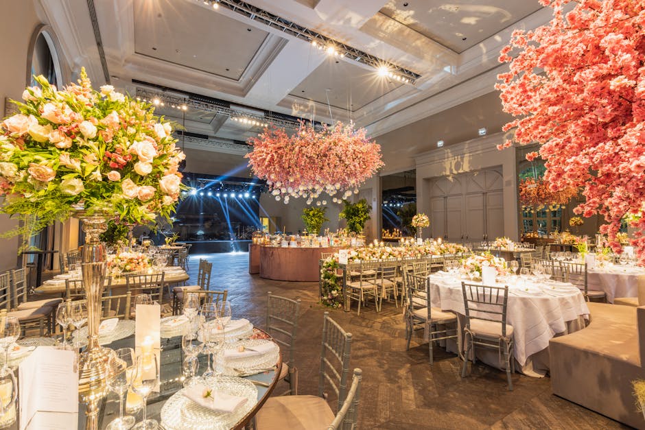 An elegant indoor wedding reception venue decorated with large floral arrangements featuring light pink and cream roses, peonies, and greenery in tall, silver vases. The arrangement includes a central