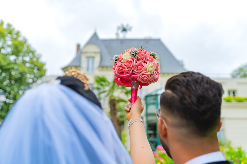 A person holding a round floral bouquet arrangement composed of pink and cream roses with green foliage accents, wrapped with pink ribbon, in an outdoor setting. The bouquet is positioned in front of 