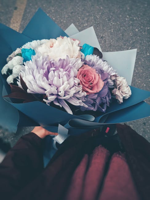 A hand holds a beautifully arranged bouquet of fresh flowers wrapped in blue and white paper, featuring large purple chrysanthemums, a single pink rose, white baby's breath, and small bright blue acce